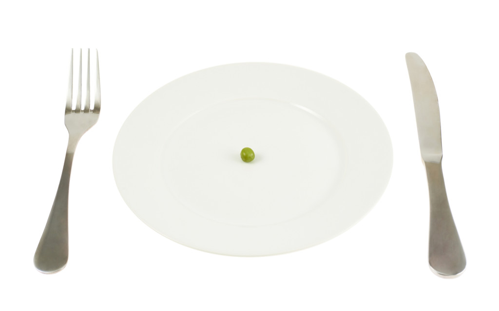Knife and fork next to a plate with a single green pea isolated over white background